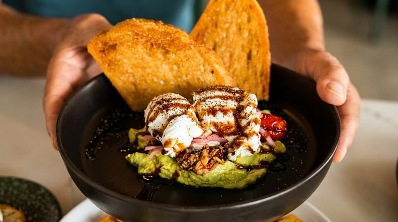 Hands present a black bowl of mashed avocado, poached eggs, tomatoes, balsamic glaze, and toasted bread.