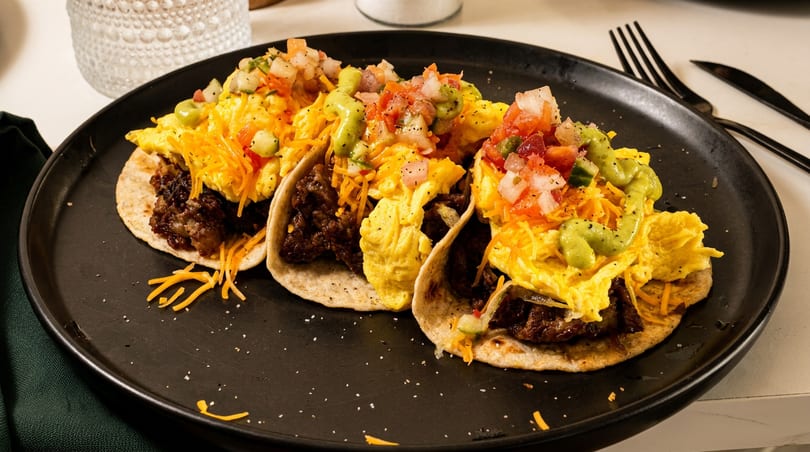 Three breakfast tacos filled with meat, scrambled eggs, cheese, pico de gallo, and avocado crema.