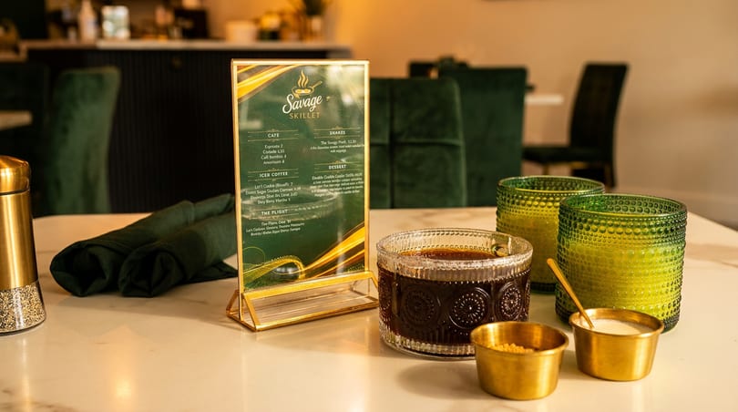 Savage Skillet menu, dark drink, green glasses, and brass condiment bowls on a restaurant table.